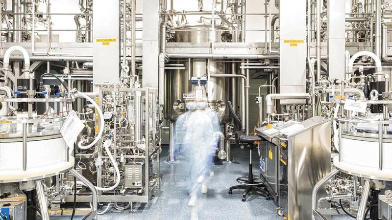 Un opérateur de production de phase, en tenue de protection, sa silhouette est floutée, au milieu d'équipements de bioproduction, cuves et tuyaux en inox.