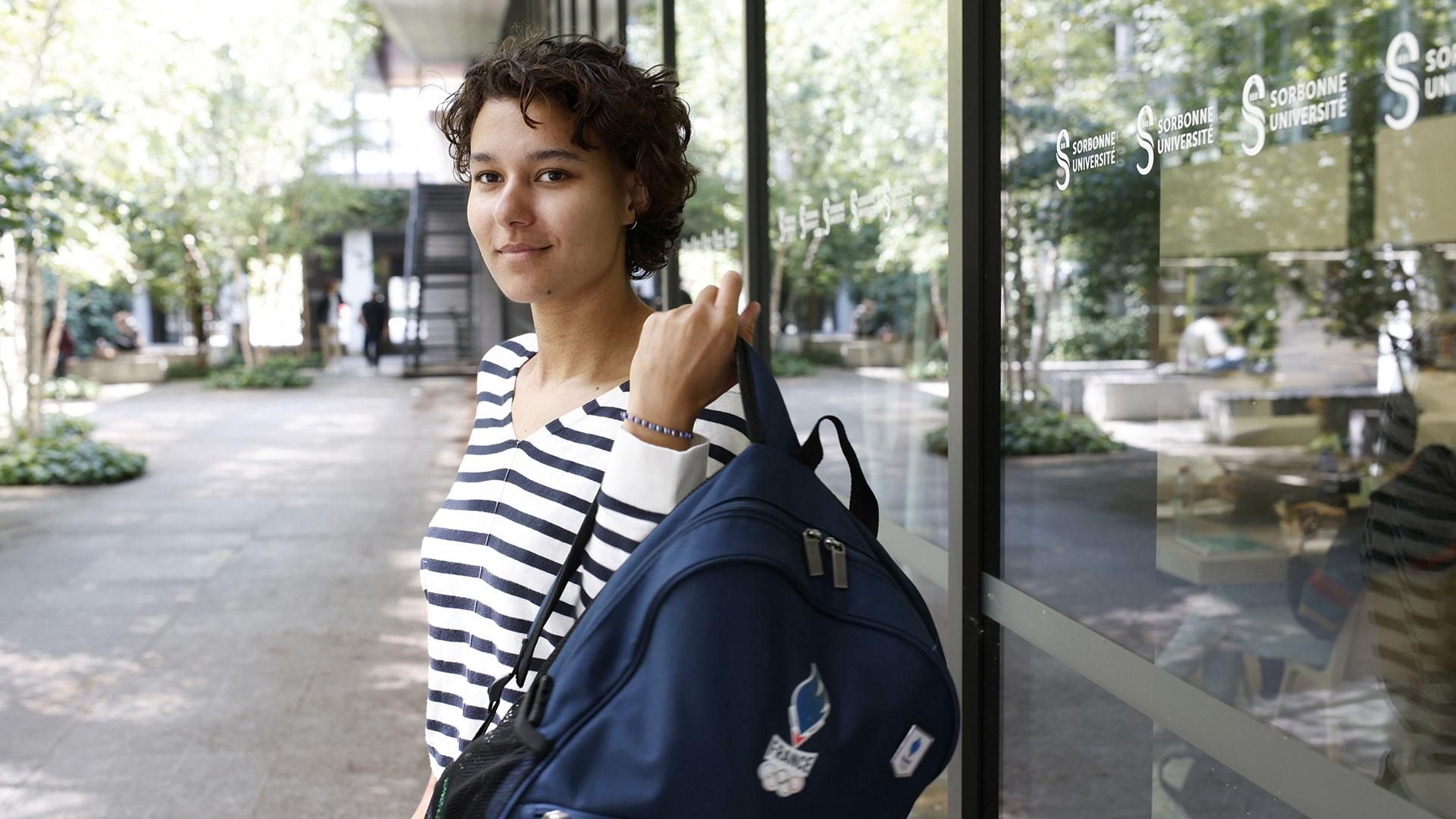 Eloïse, étudiante souriante avec un sac à dos devant un bâtiment universitaire.