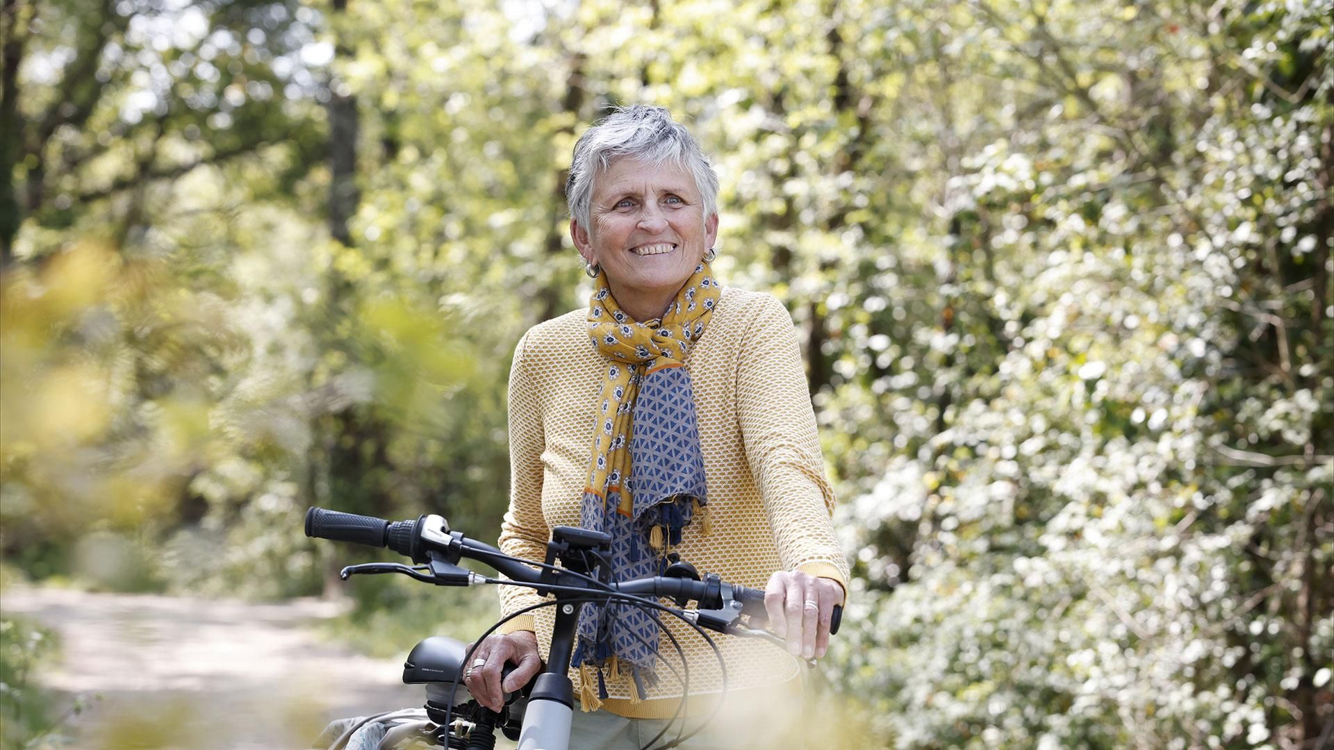 Chantal en pull jaune et écharpe bleue à motifs, souriante, tenant le guidon de son vélo dans un environnement naturel.