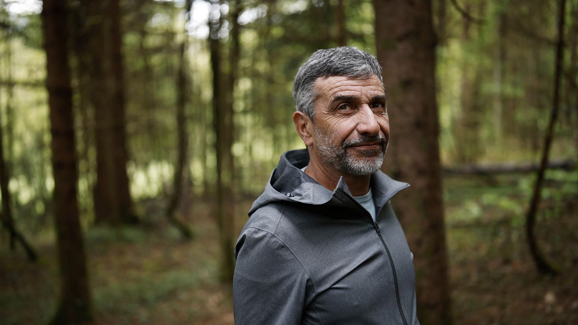 Franck en veste grise à capuche, souriant paisiblement dans une forêt dense et verdoyante.