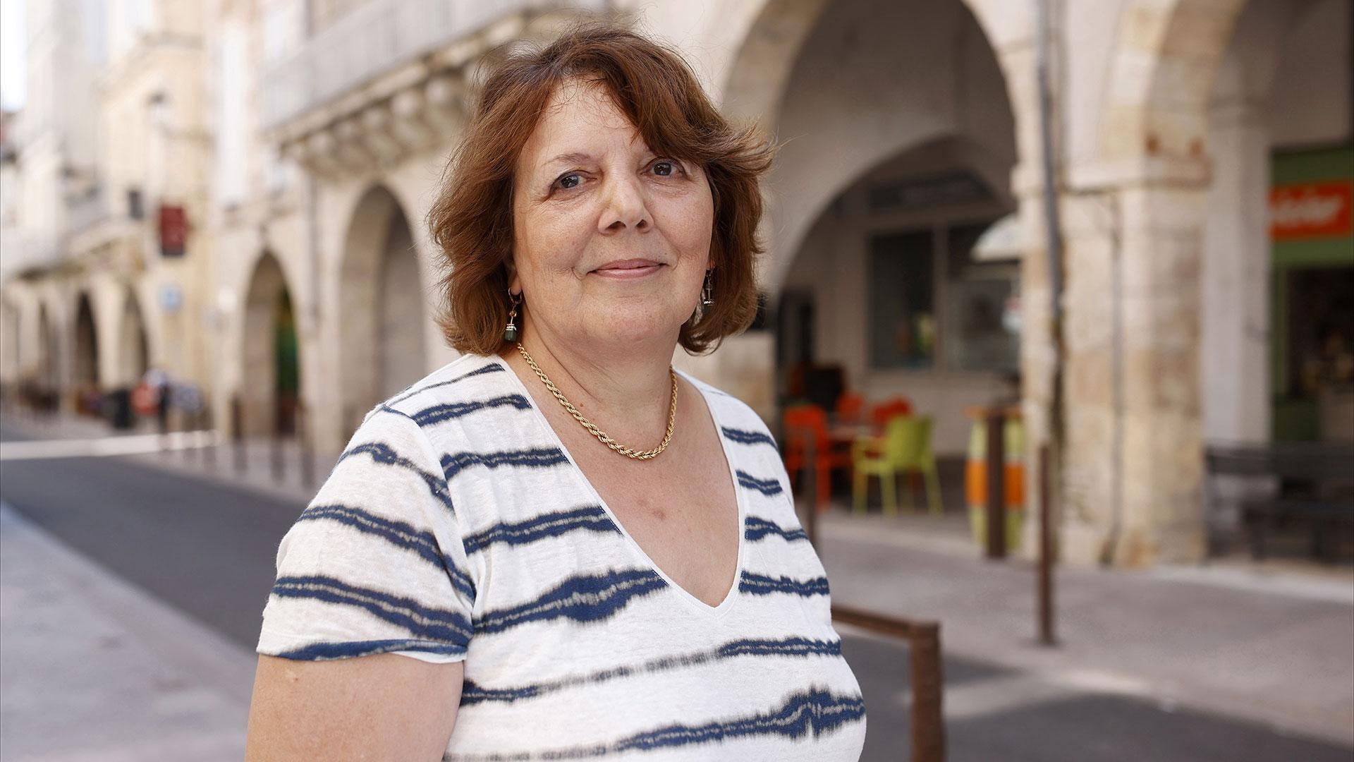 Christine en top rayé blanc et bleu, souriante, debout dans une rue avec terrasses de café en arrière-plan.