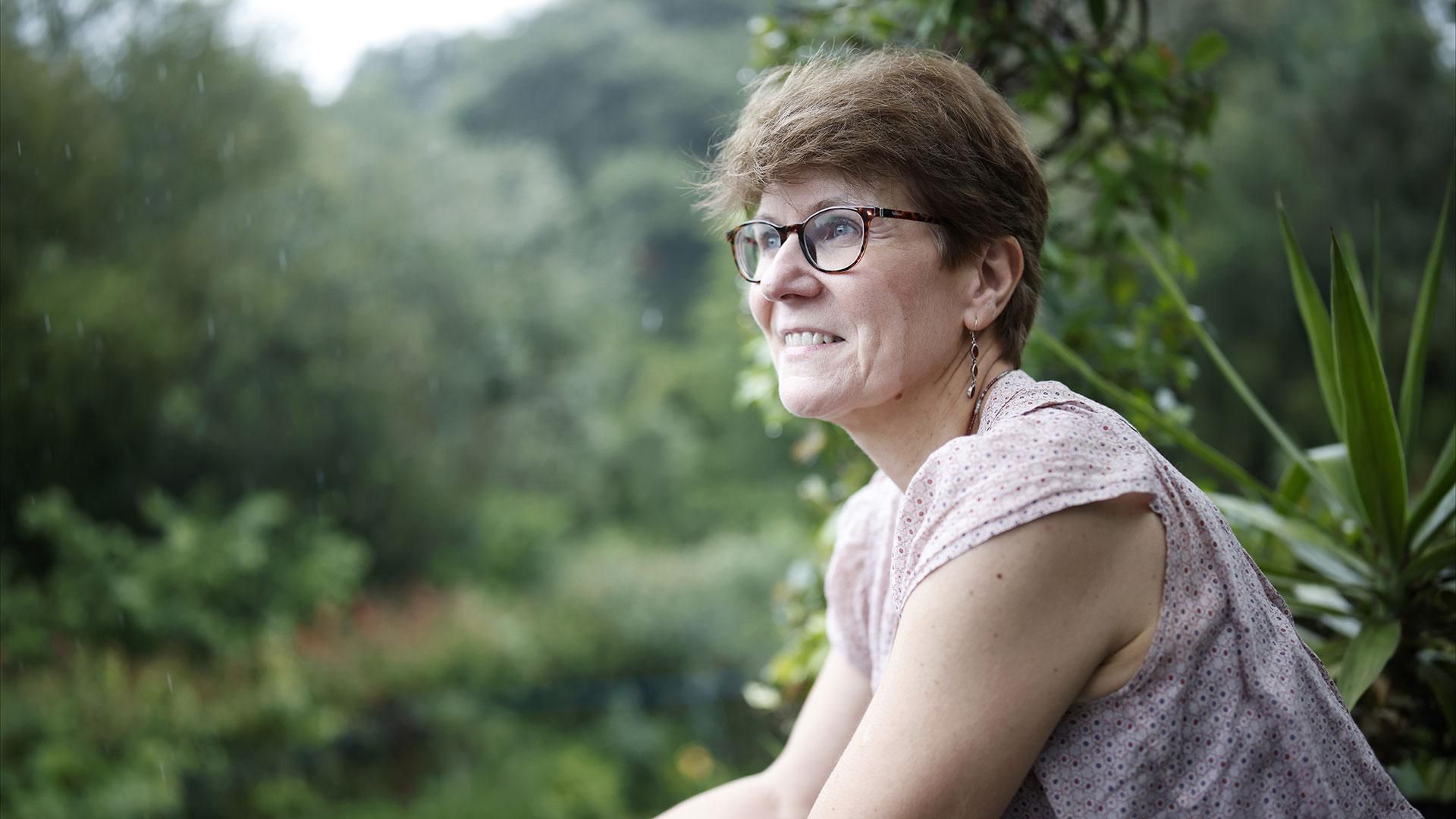 Kathy avec lunettes et chemisier à motifs, souriante dans un jardin luxuriant sous la pluie.