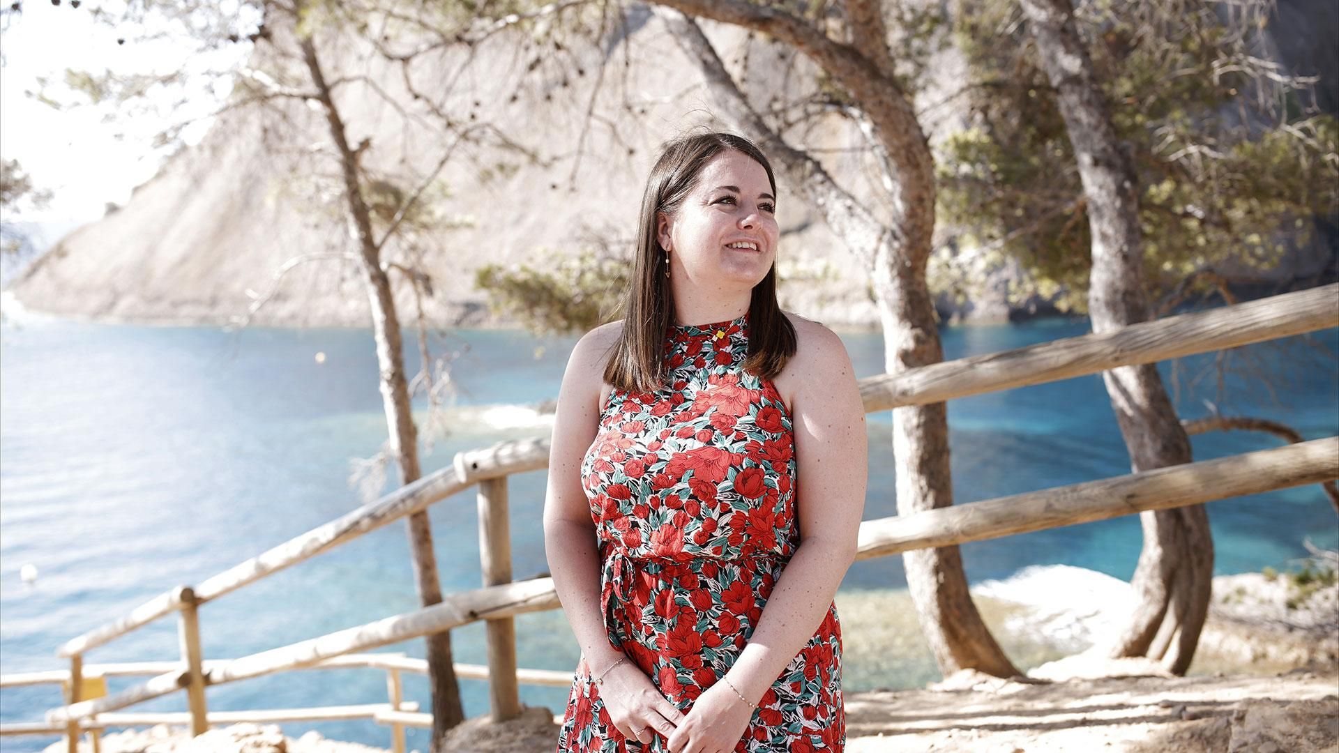 Gwenaëlle en robe rouge à motifs floraux, souriante sur un ponton en bois au bord d'un lac avec montagne en arrière-plan.