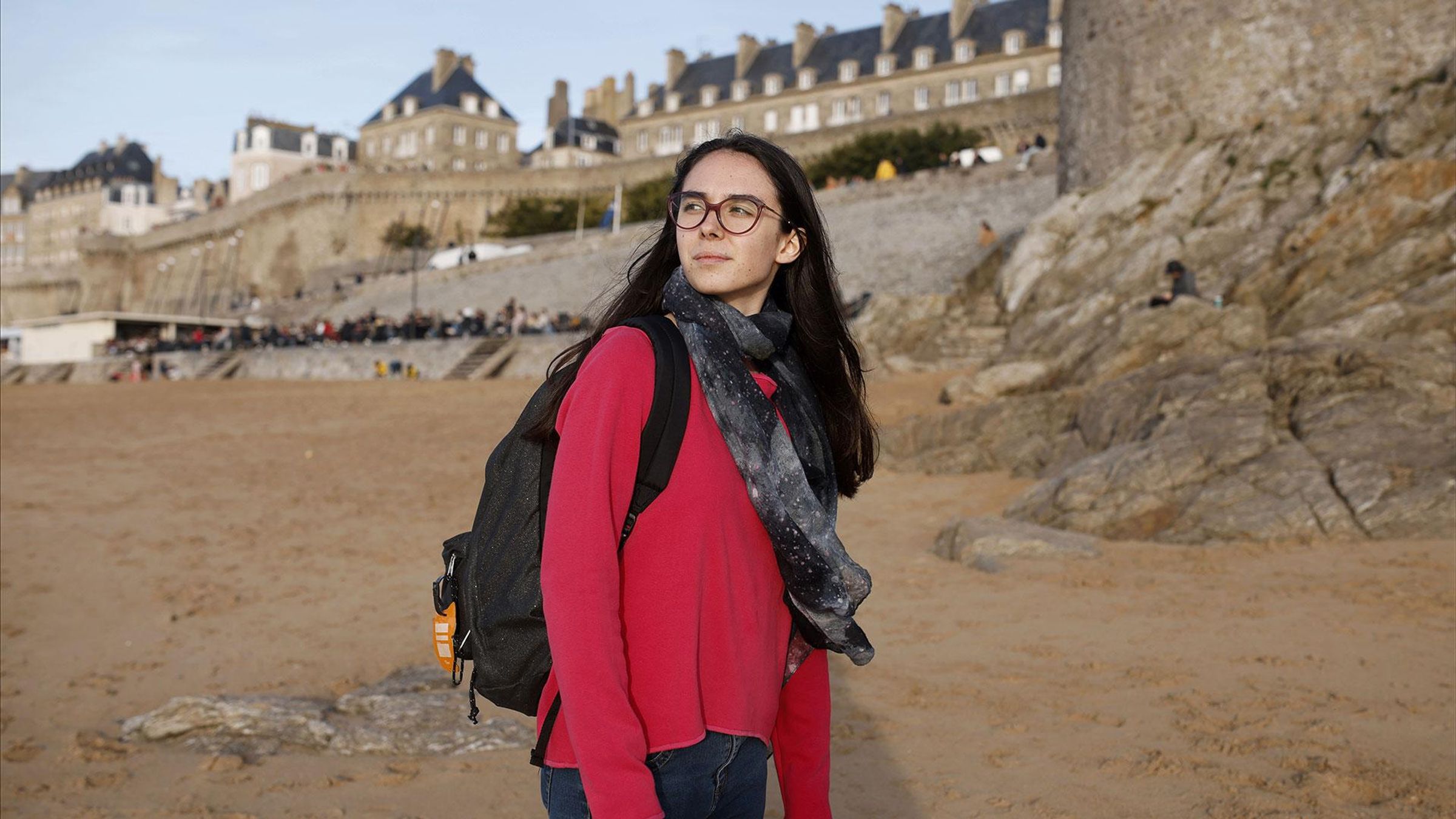 Lou en chemise rouge et écharpe, regardant vers l'horizon sur une plage avec côte rocheuse.