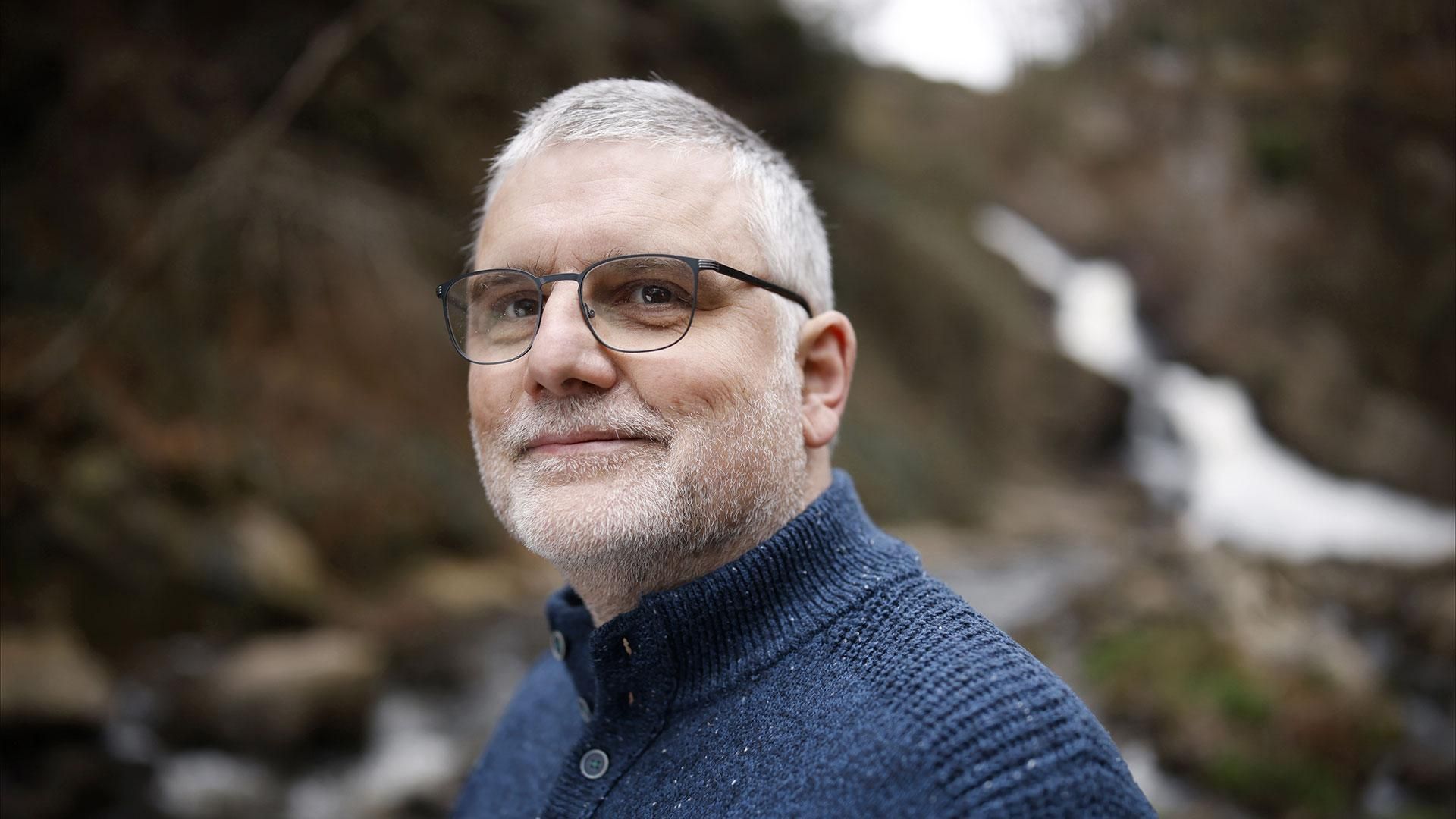 Bernard aux cheveux et barbe blancs, portant des lunettes et un pull bleu marine, souriant.