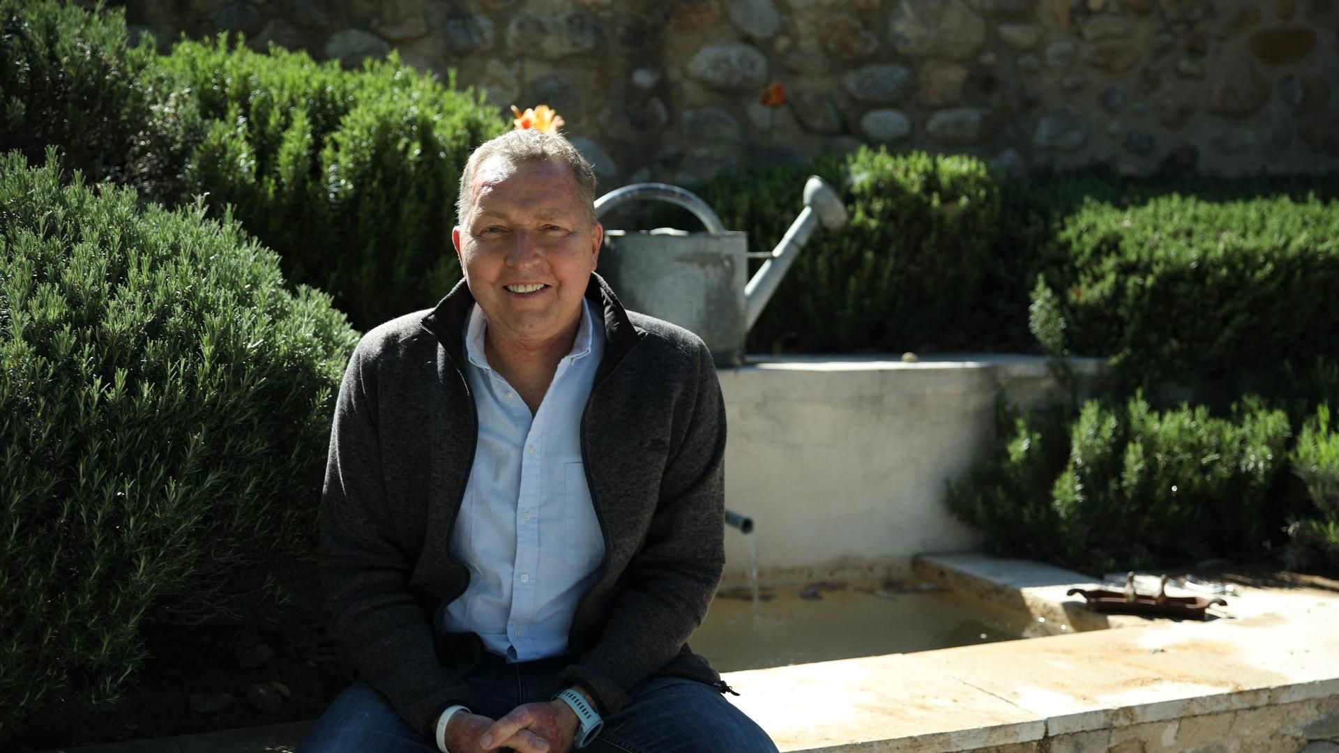 Benjamin en chemise bleue et gilet gris, souriant, assis sur un muret de pierre dans un jardin verdoyant